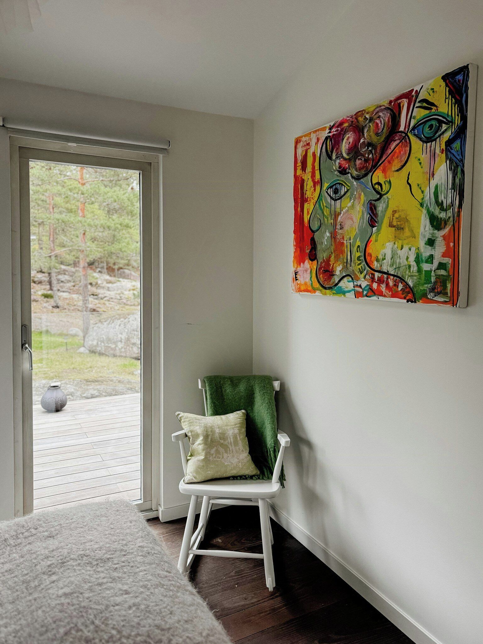 Une pièce avec une chaise blanche, un plaid vert, un tableau abstrait aux couleurs vives et une vue à travers une porte vitrée.