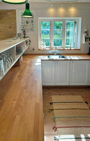 A kitchen with a wooden floor, white cabinets, a green pendant light, and a window with a view of greenery. - Family house with large terrace near Skødshoved (Knebel)