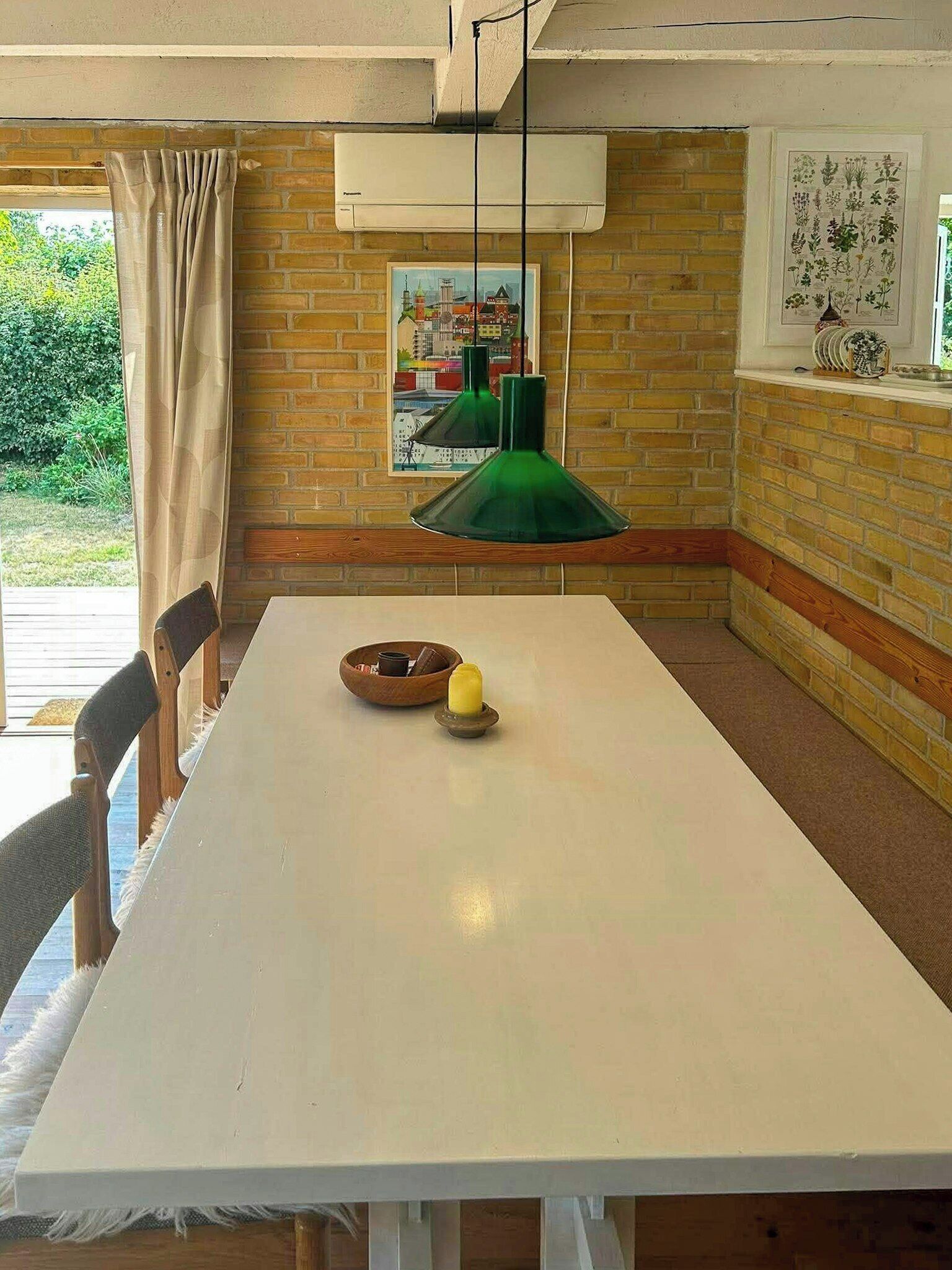 Un coin repas avec une table blanche, des chaises, une suspension verte et un mur en briques orné d’un tableau représentant un paysage urbain.