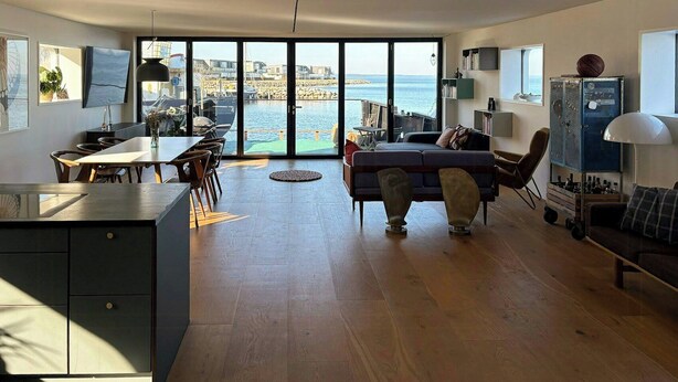 En rummelig opholdsstue med store skydedøre, der giver udsigt over havnen, et spiseområde med et træbord, og et hyggeligt siddeområde med sofa og lænestole.