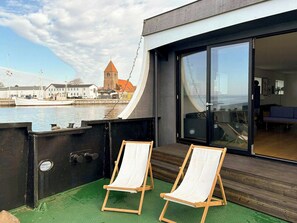 A balcony with two wooden chairs, a view of the water, and a building with a large glass door. - Converted island ferry with sea access in Stege (Stege)