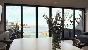 Et moderne værelse med et stort vindue, der giver udsigt over vandet, et hvidt bord og en vase med tørrede planter.