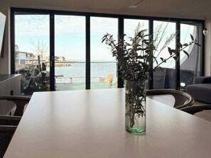 A modern room with a large window overlooking a body of water, a white table, and a vase with dried plants. - Converted island ferry with sea access in Stege (Stege)