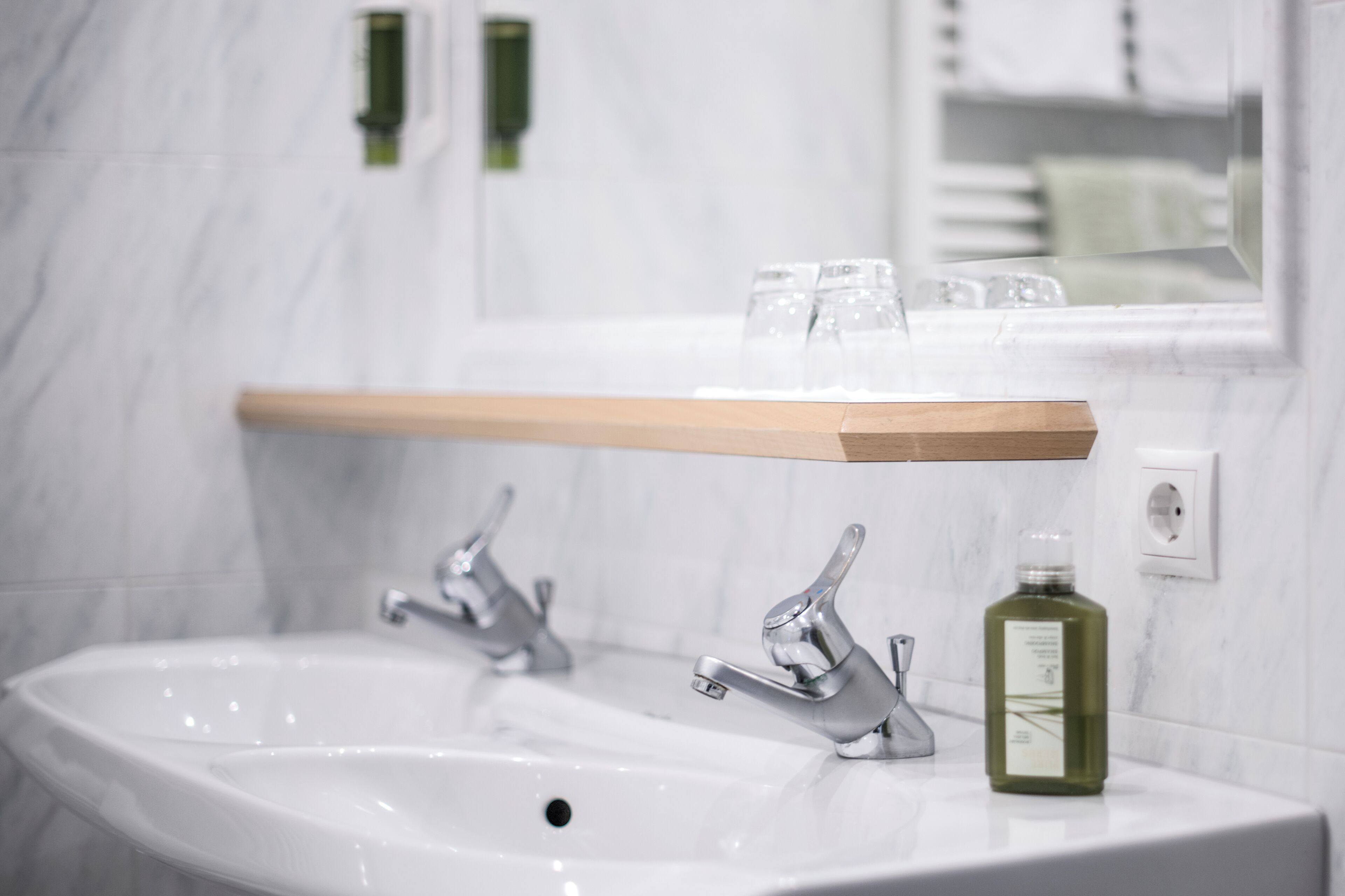 A bathroom with a white sink, two faucets, a wooden shelf above the sink, and a bottle of liquid soap.