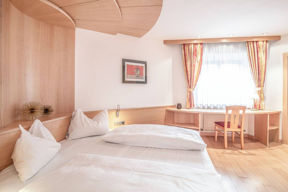 Habitación de hotel con cama, escritorio, silla, un cuadro en la pared y ventana con cortinas.