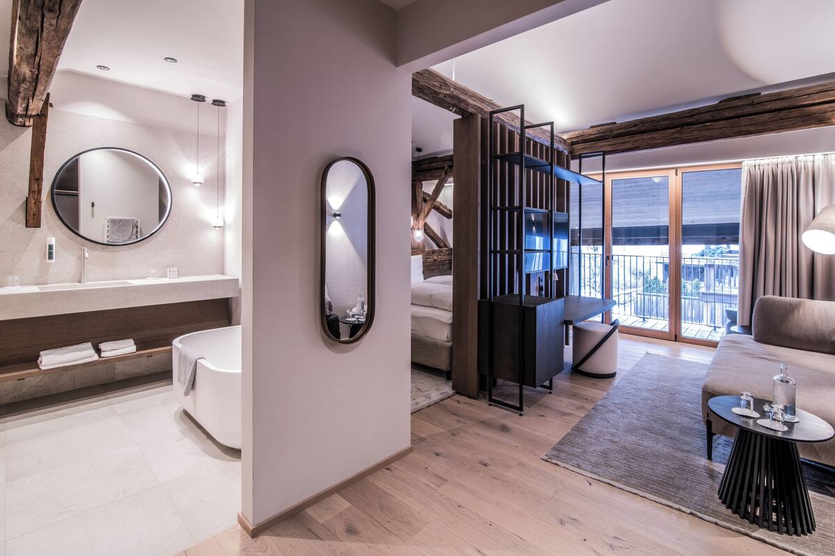Habitación de hotel moderna con baño, un espejo grande, sofá, mesa de comedor y balcón con vistas.