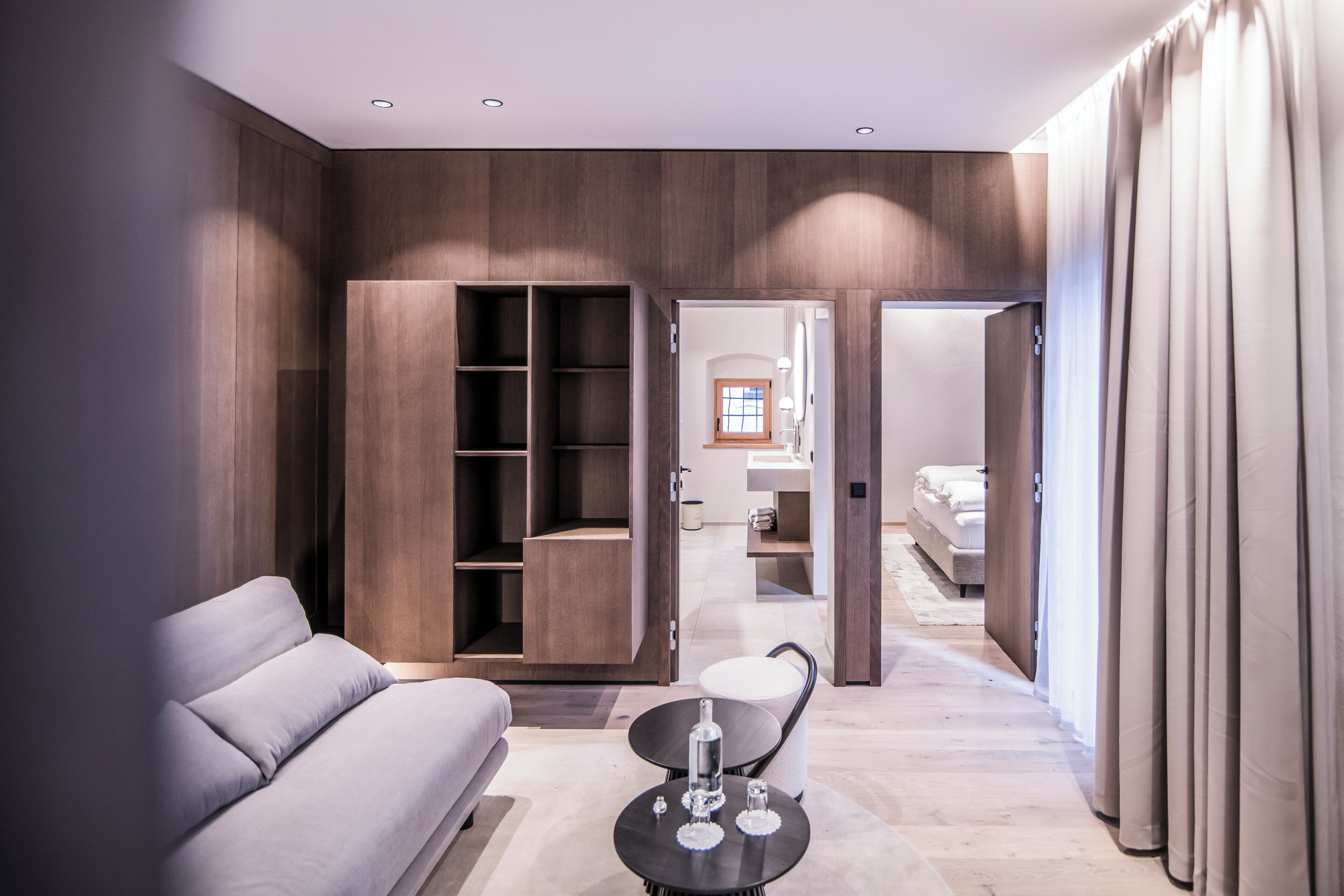 A modern hotel room with a sofa, a small round table, a built-in wardrobe, and a connecting door to a bathroom.