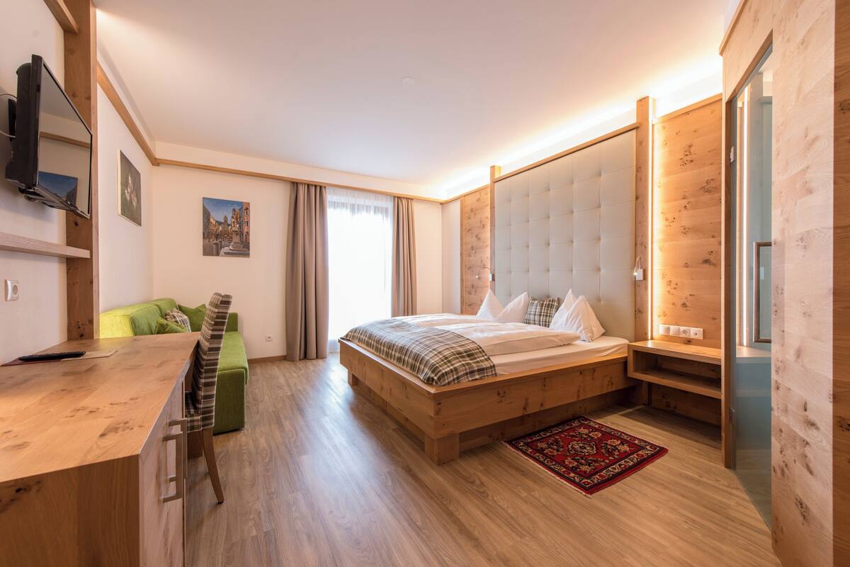 Un dormitorio moderno con una cama de madera, un escritorio, un sofá verde y un amplio ventanal con cortinas.