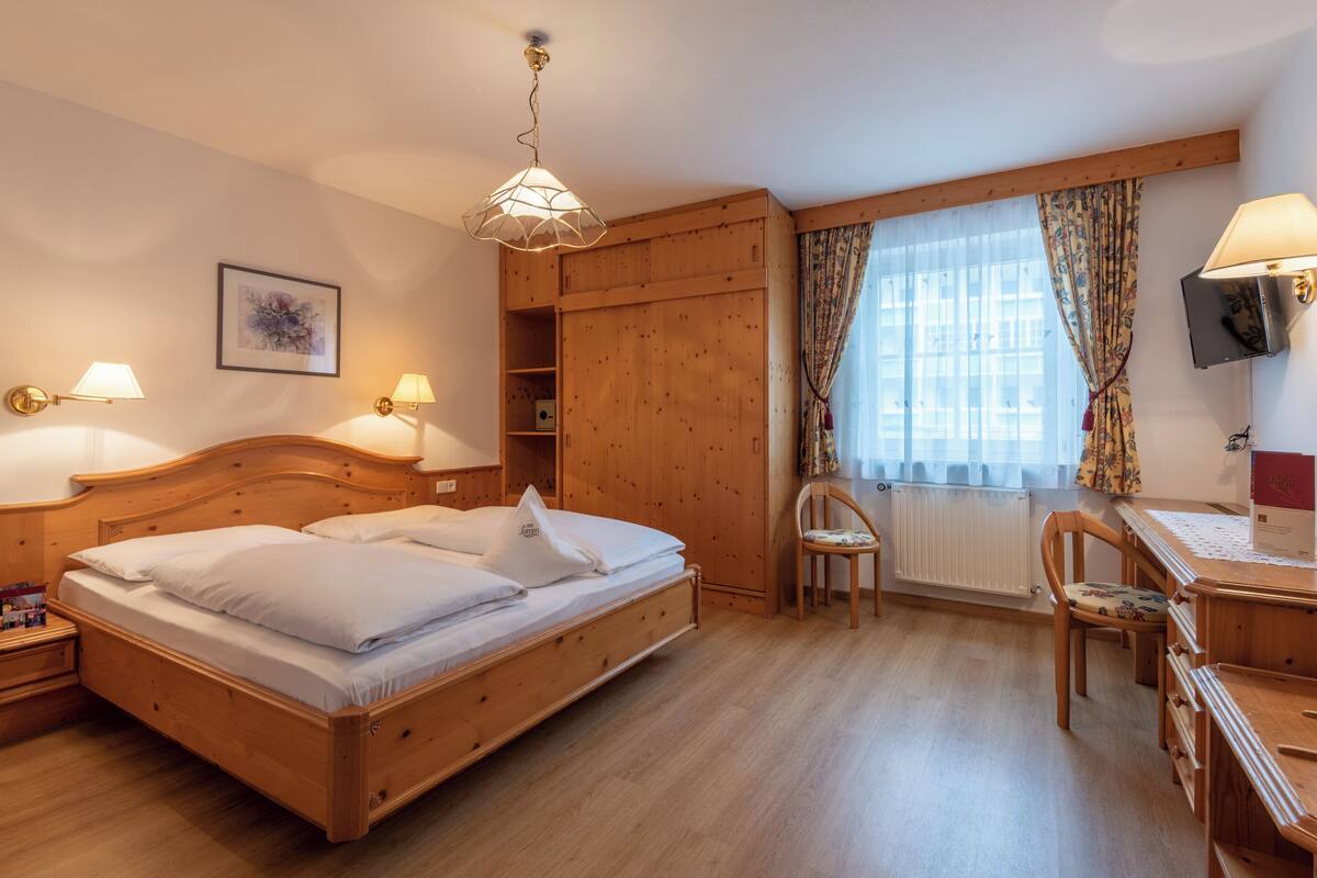 Una cama de madera con cabecera, lámparas de noche, un escritorio con silla y una ventana con cortinas.