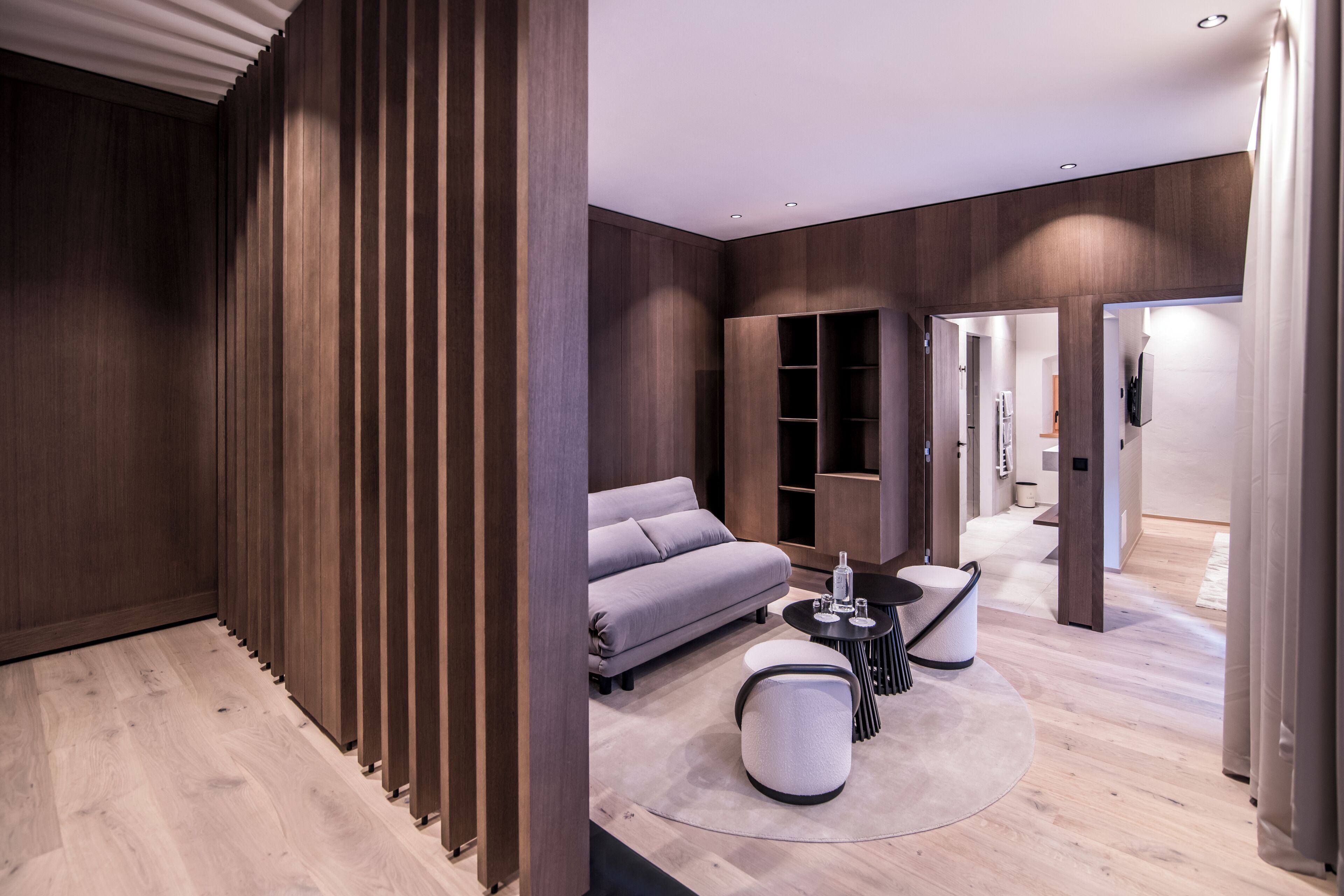 A modern interior with a sofa, round coffee table, and ottoman, wooden paneling, and a view into another room.