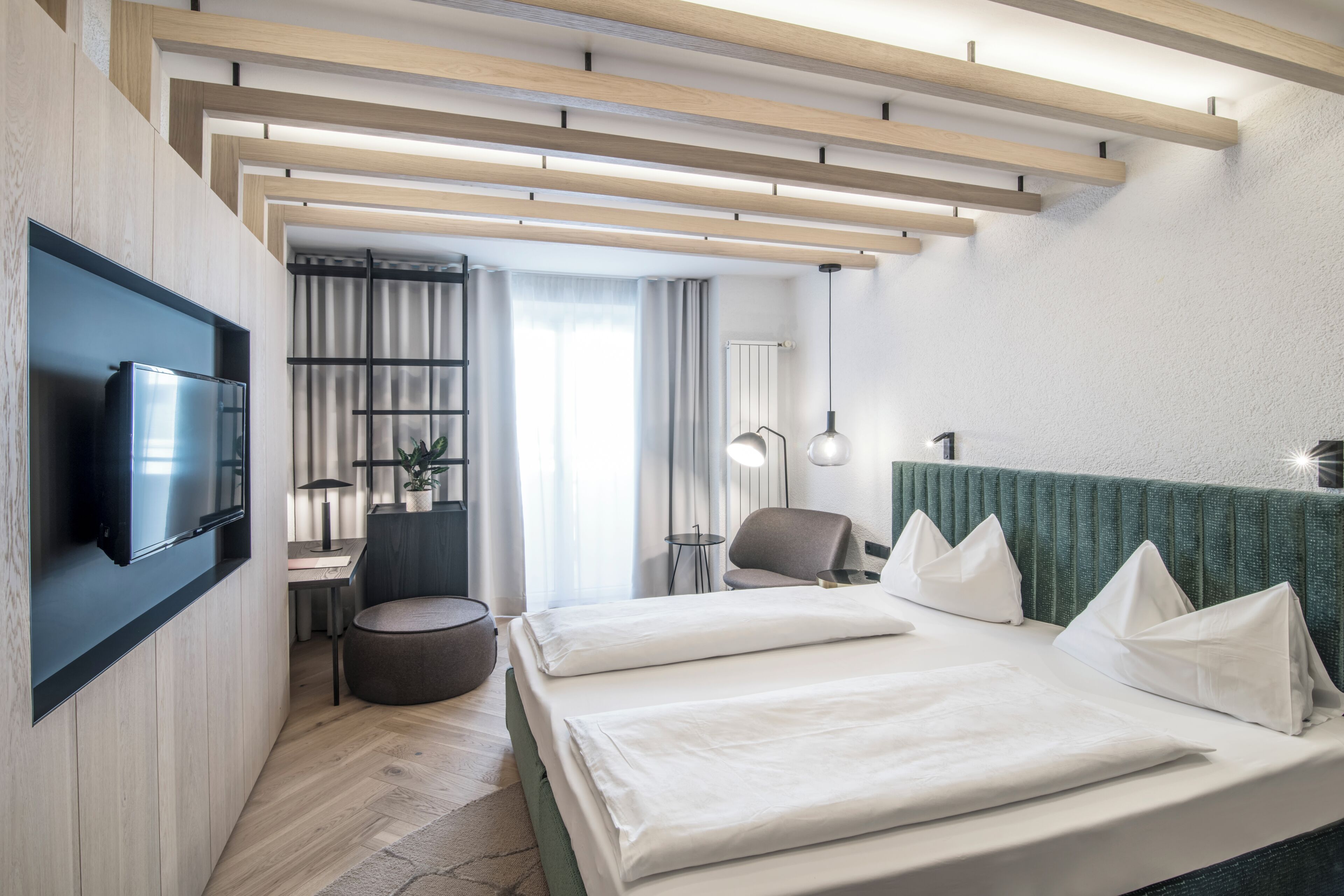 Una camera d'albergo moderna con TV a schermo piatto, un letto con lenzuola bianche, una scrivania con sedia, una lampada e una parete verde decorativa.
