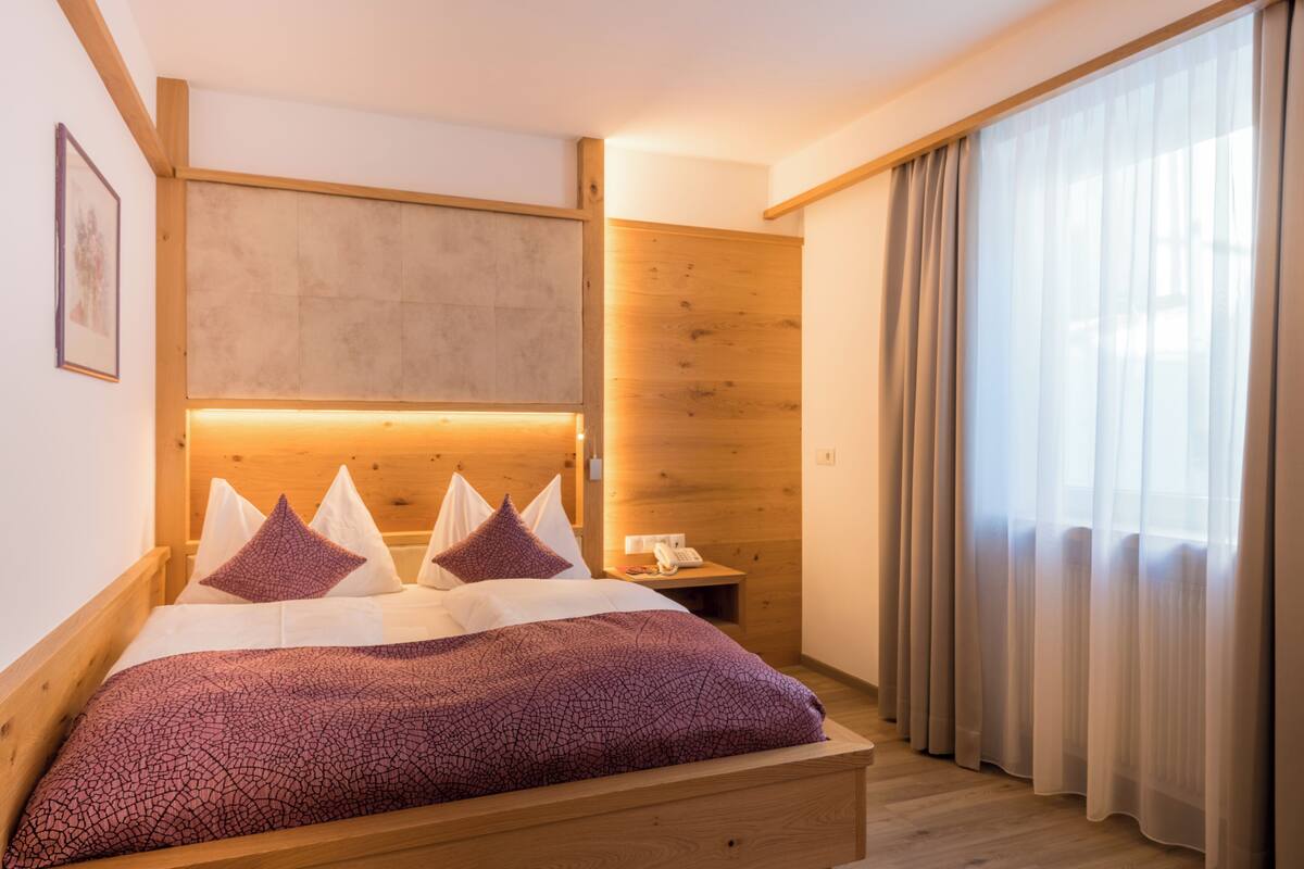 Habitación de hotel con cama de madera, una mesita de noche, teléfono y un ventanal grande con cortinas.