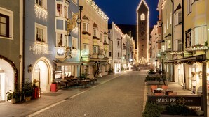 A street lined with buildings, some with illuminated signs, and a clock tower in the distance.