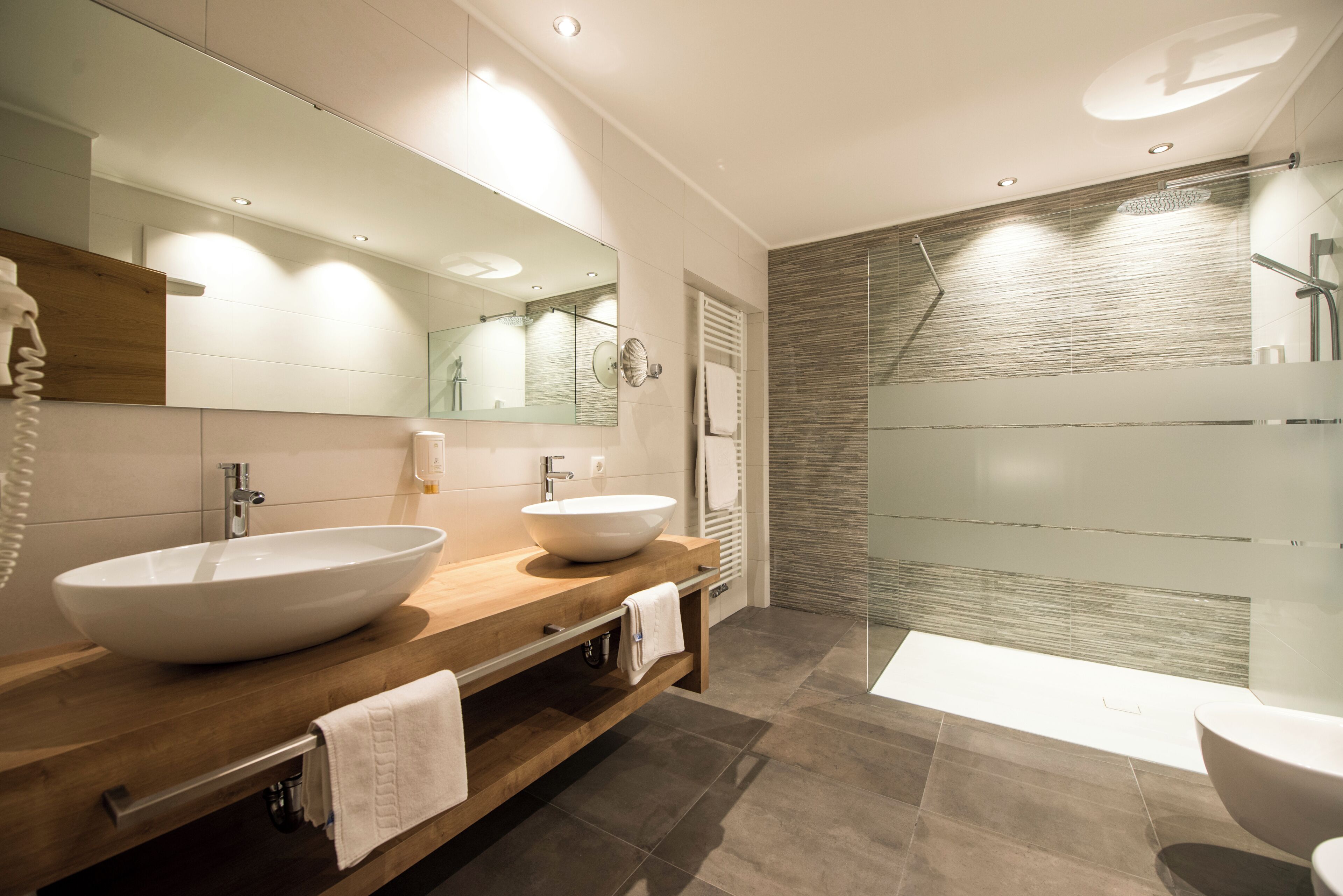 Un baño moderno con tocador de madera, dos lavamanos, un espejo grande y cabina de ducha con paredes de vidrio.