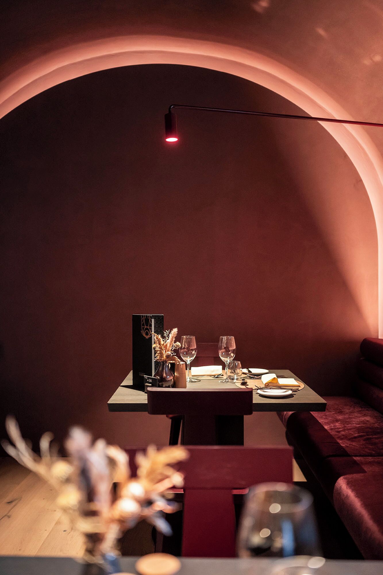 Un comedor tenuemente iluminado con una mesa dispuesta para dos, un jarrón con flores secas y un sofá rojo.