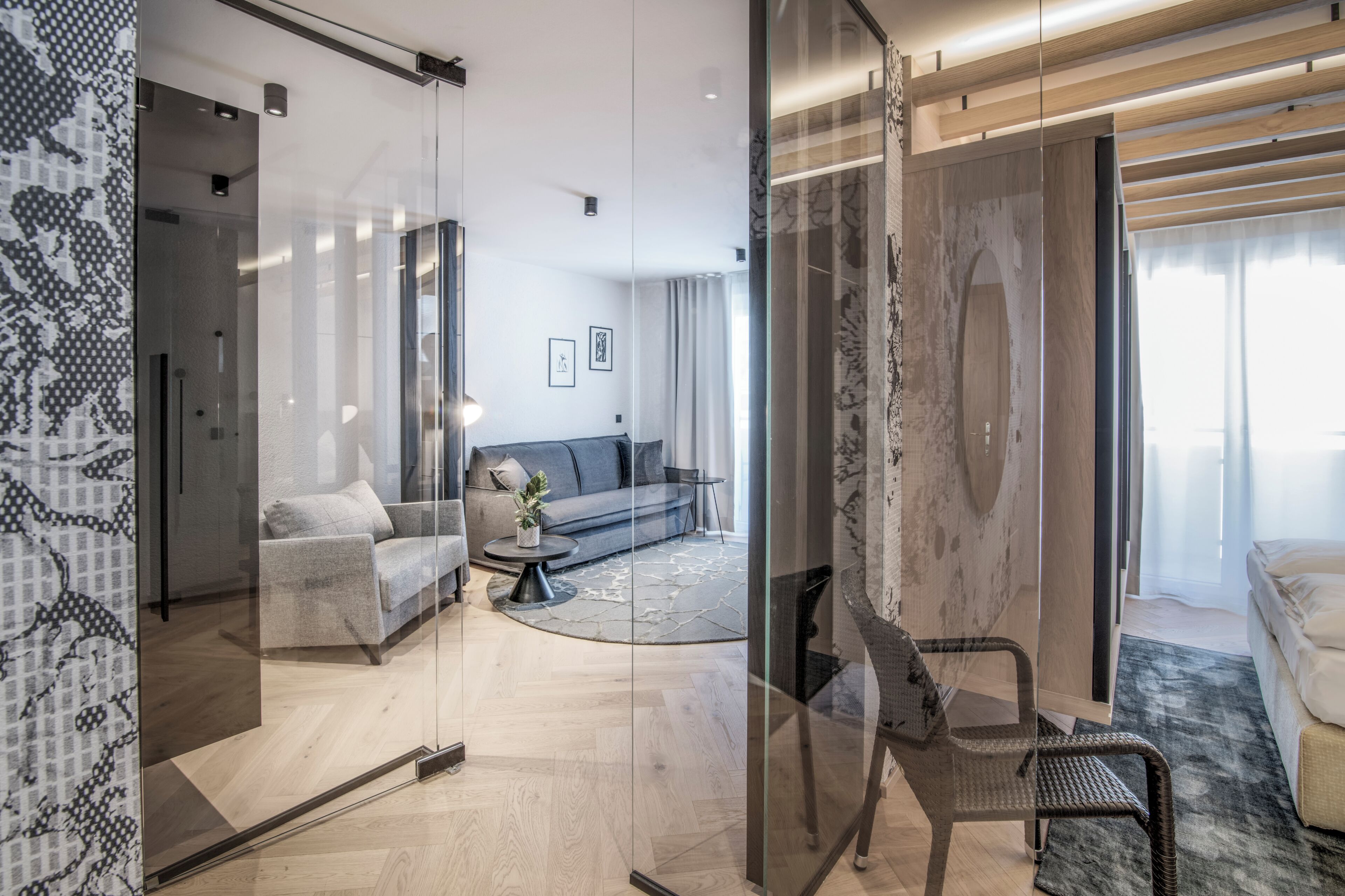 A modern hotel room with a glass partition, a sofa, a chair, and a bed.