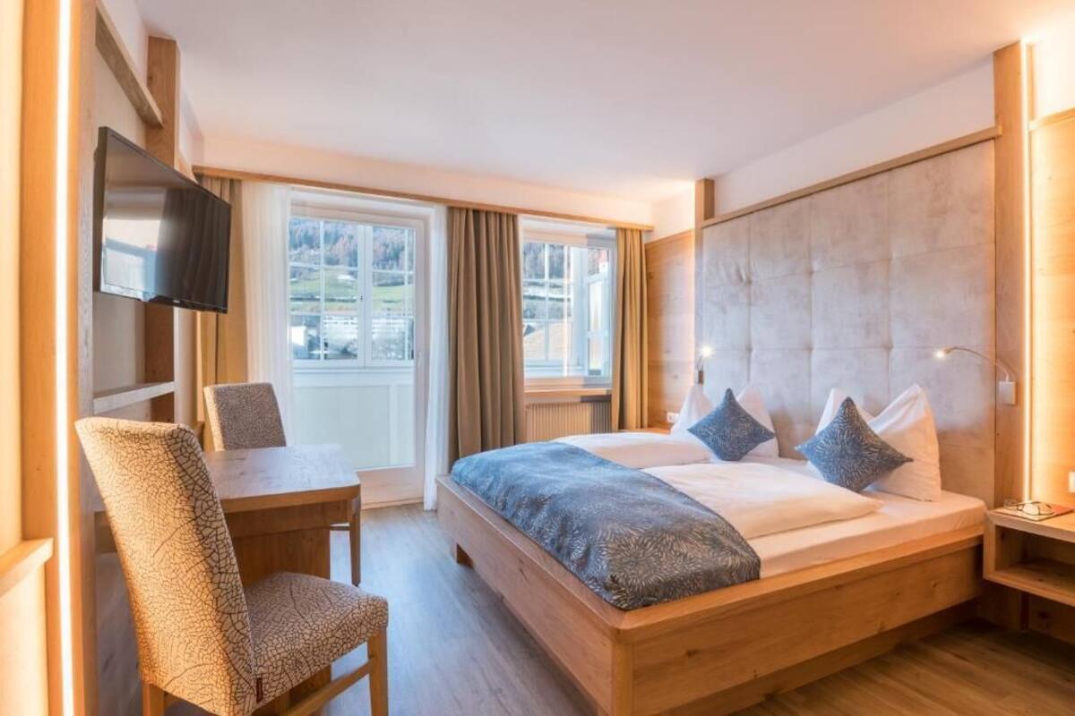 Un dormitorio con cama, escritorio, silla, televisor y ventana con cortinas.