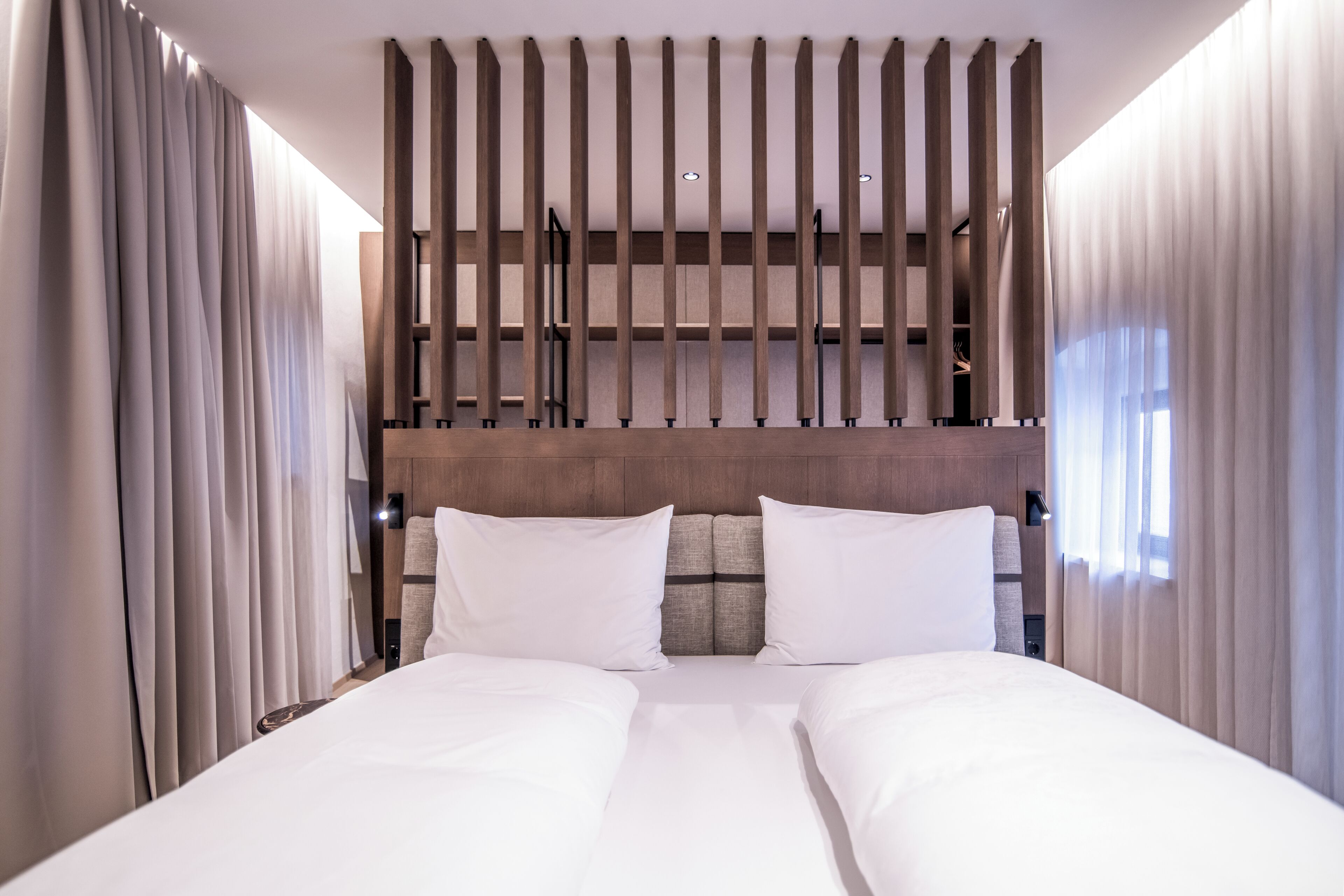 A hotel room with a large bed, curtains, a wooden headboard, and a wall-mounted shelf.