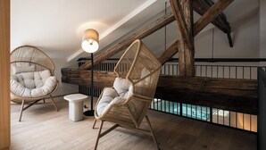 A loft-style room with exposed wooden beams, two wicker chairs with cushions, a small round table, and a hanging light fixture.
