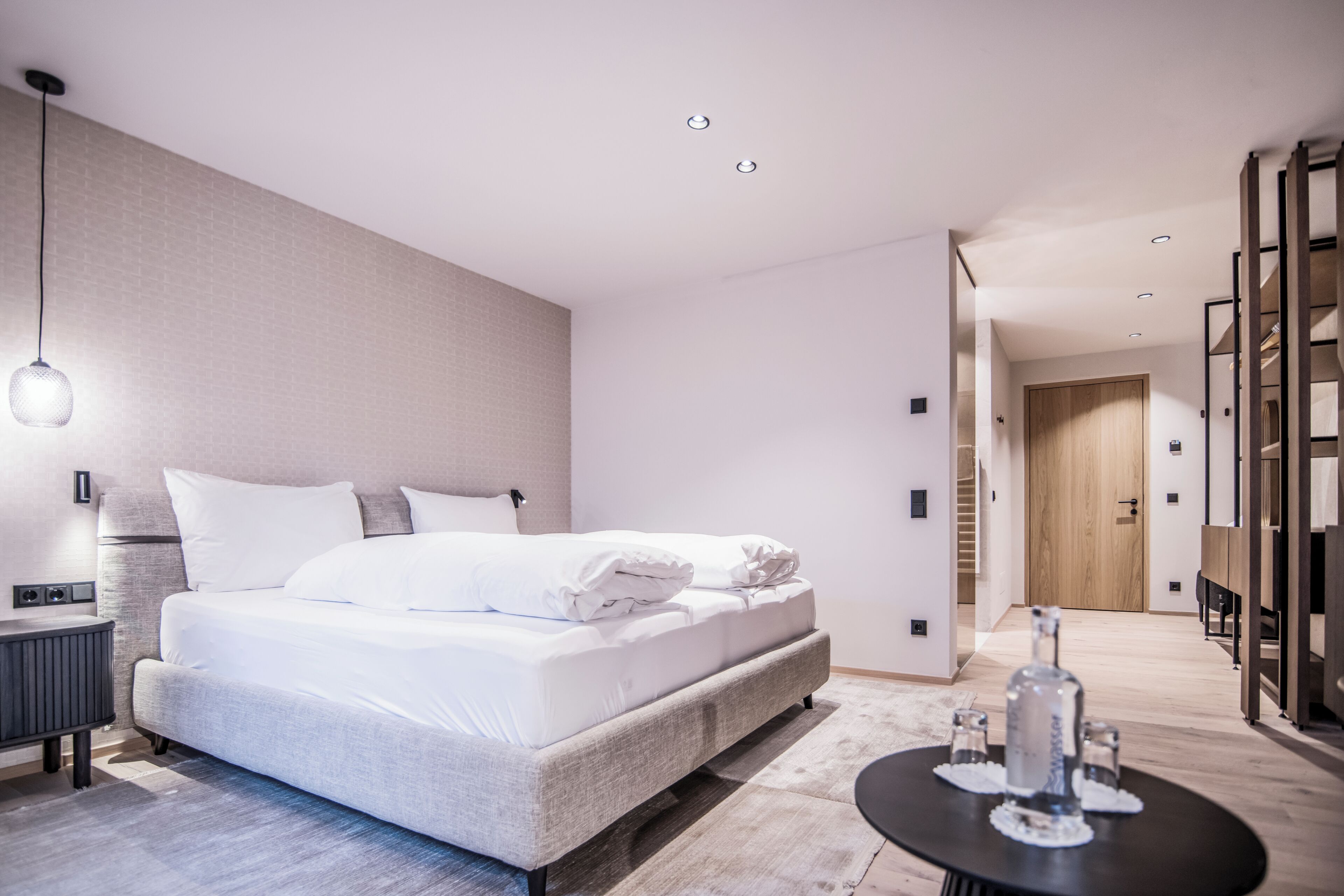 A modern hotel room with a large bed, bedside table, and a view of the hallway.