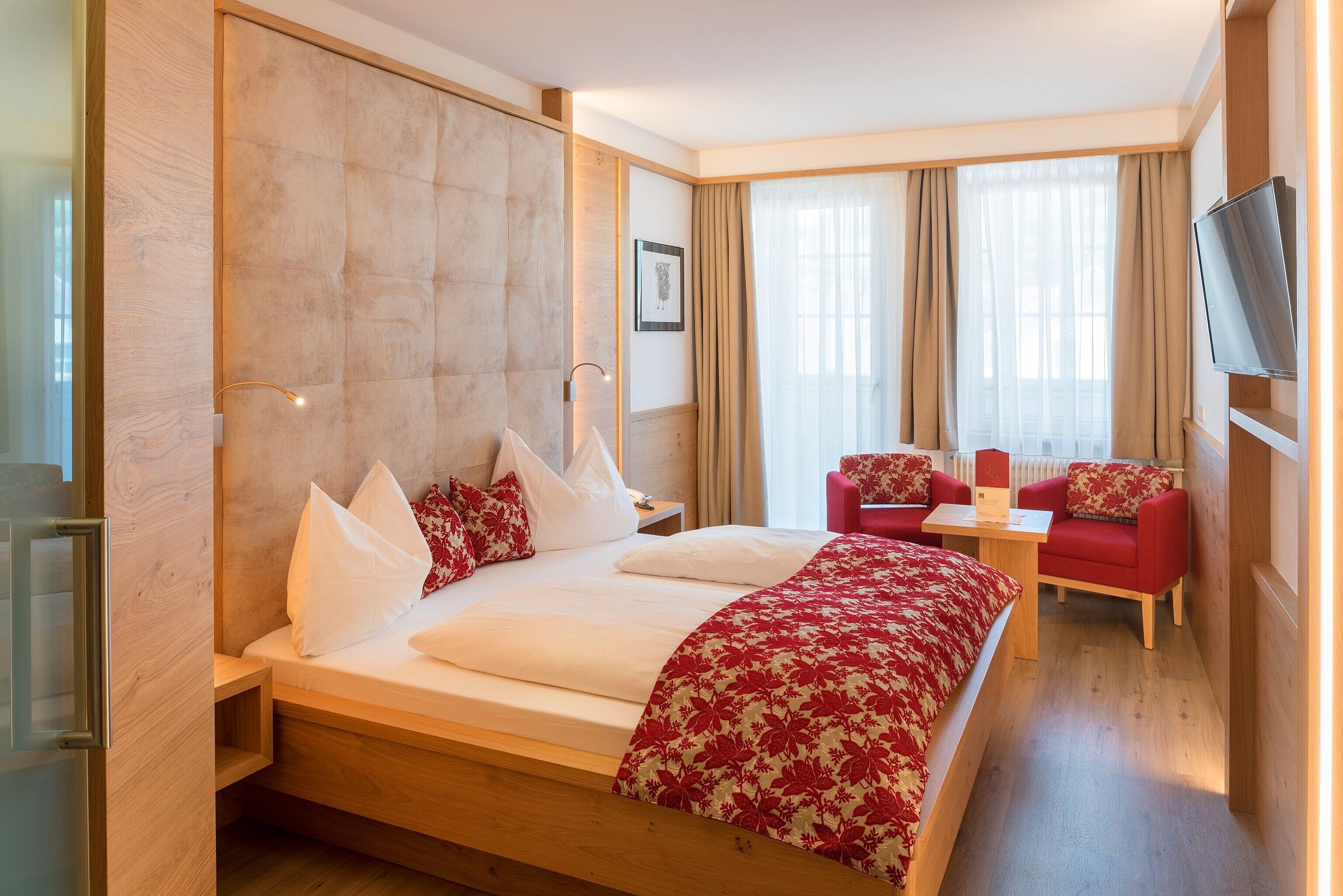 Habitación de hotel con una cama grande, un escritorio, dos sillas, un televisor y un ventanal con cortinas.