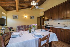 Un coin repas avec une table dressée pour quatre personnes, des meubles en bois, un ventilateur de plafond et une cuisine en arrière-plan.