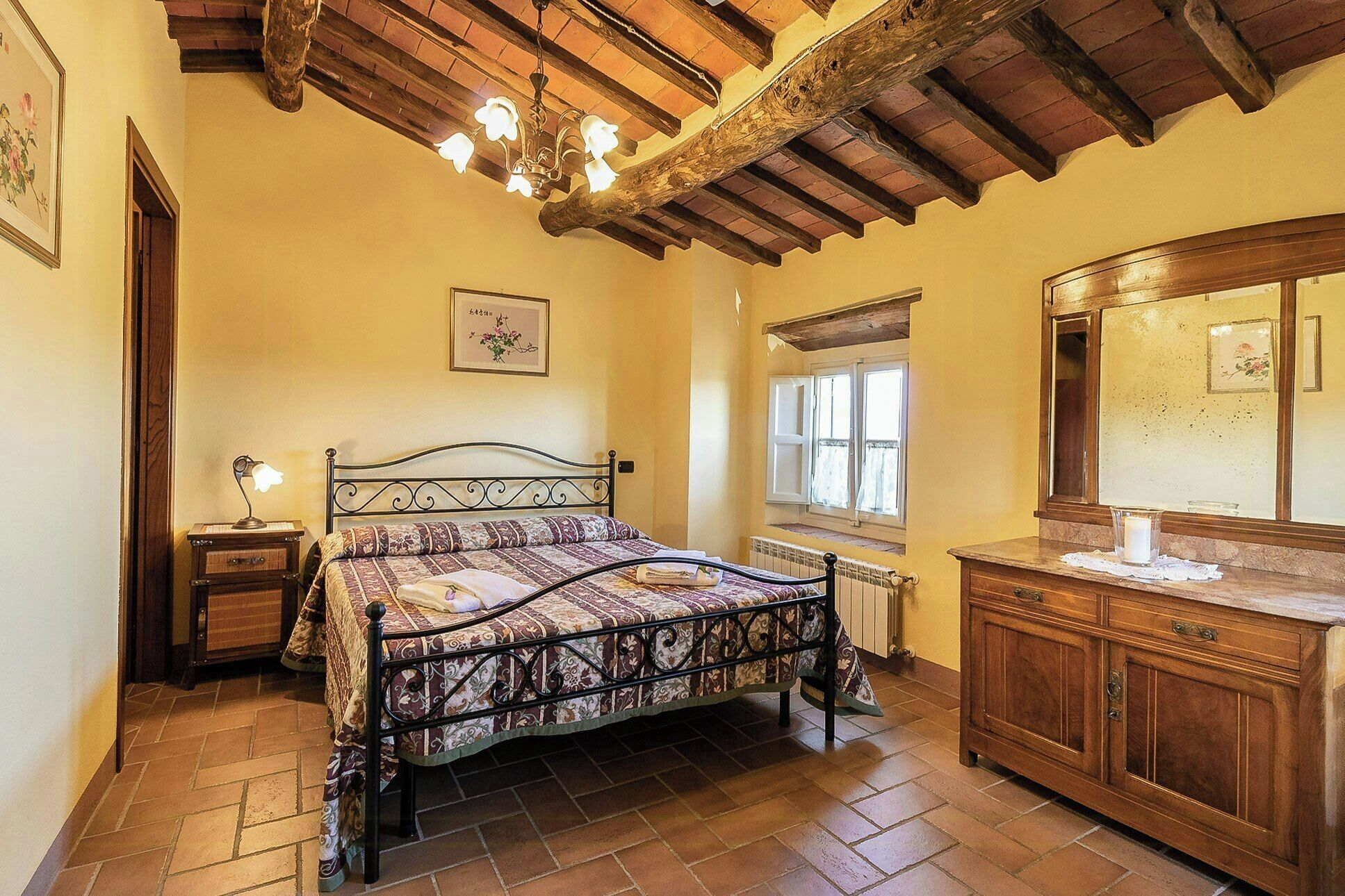 Ein Schlafzimmer mit Bett, Nachttisch, Kommode, Spiegel, Fenster und einer Chandelier.