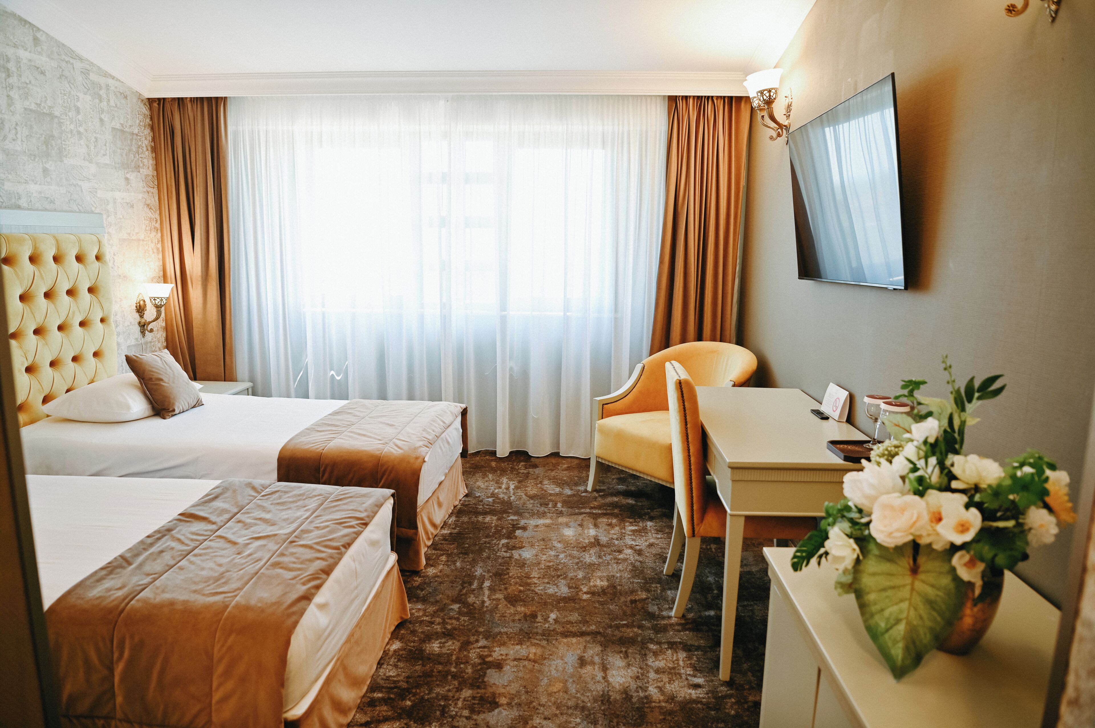 A hotel room with two beds, a desk, a chair, a TV, and a flower vase.