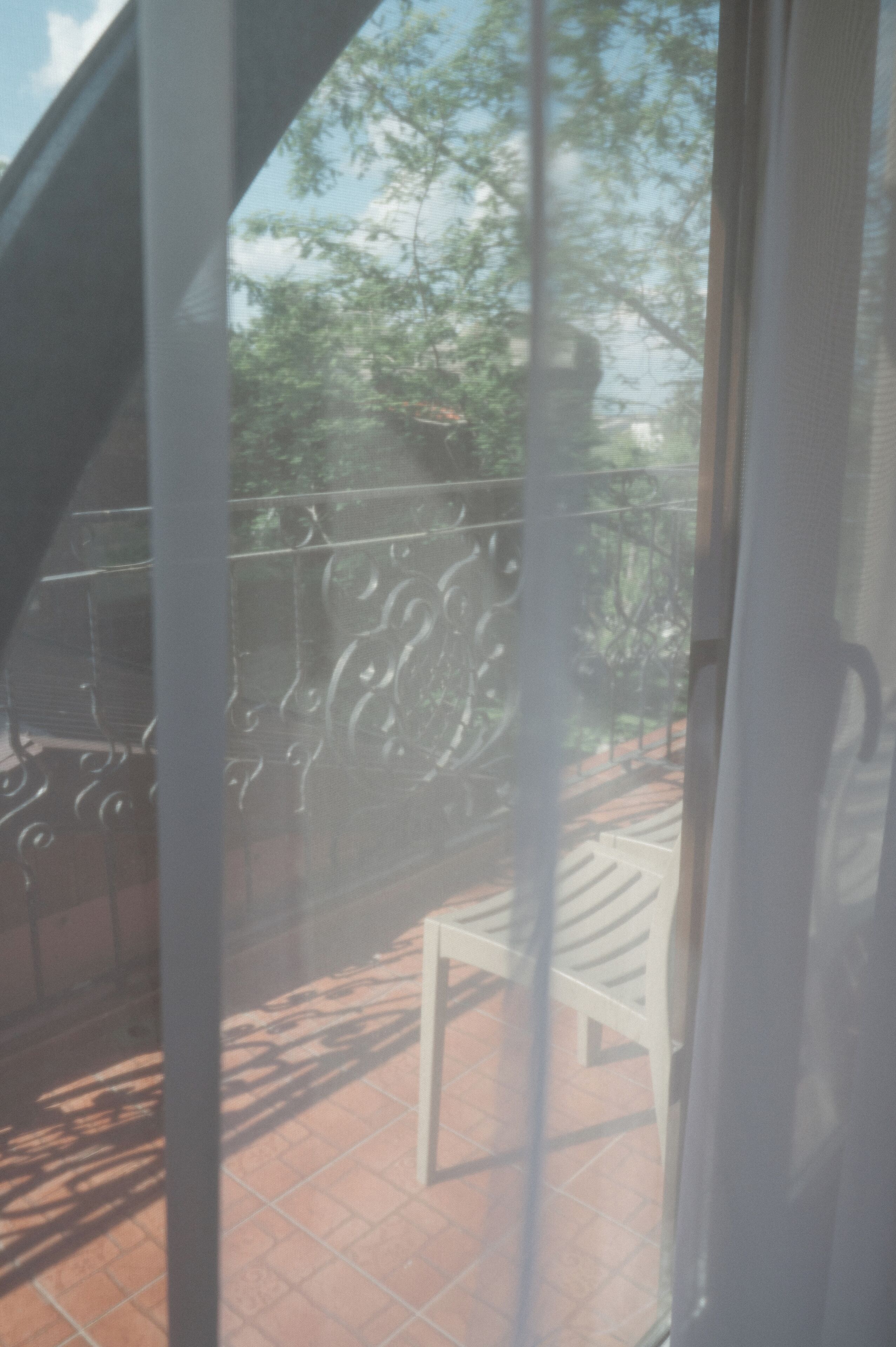 Ein Balkon mit Metallgeländer, einem weißen Stuhl und Blick auf Bäume sowie ein Gebäude im Hintergrund.
