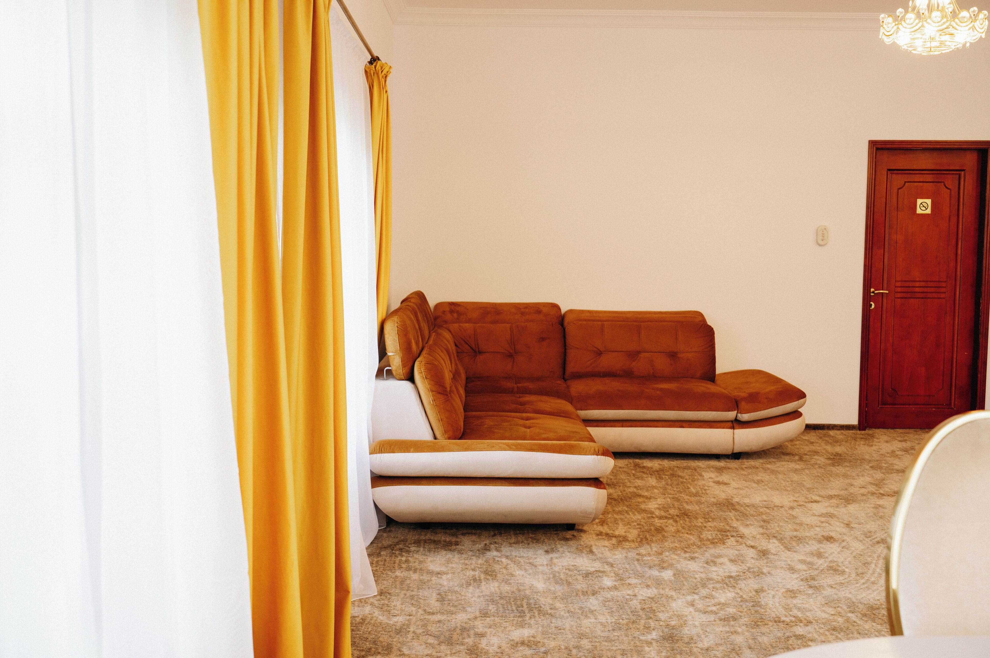 A room with a brown sectional sofa, a white chair, a wooden door, and a yellow curtain.