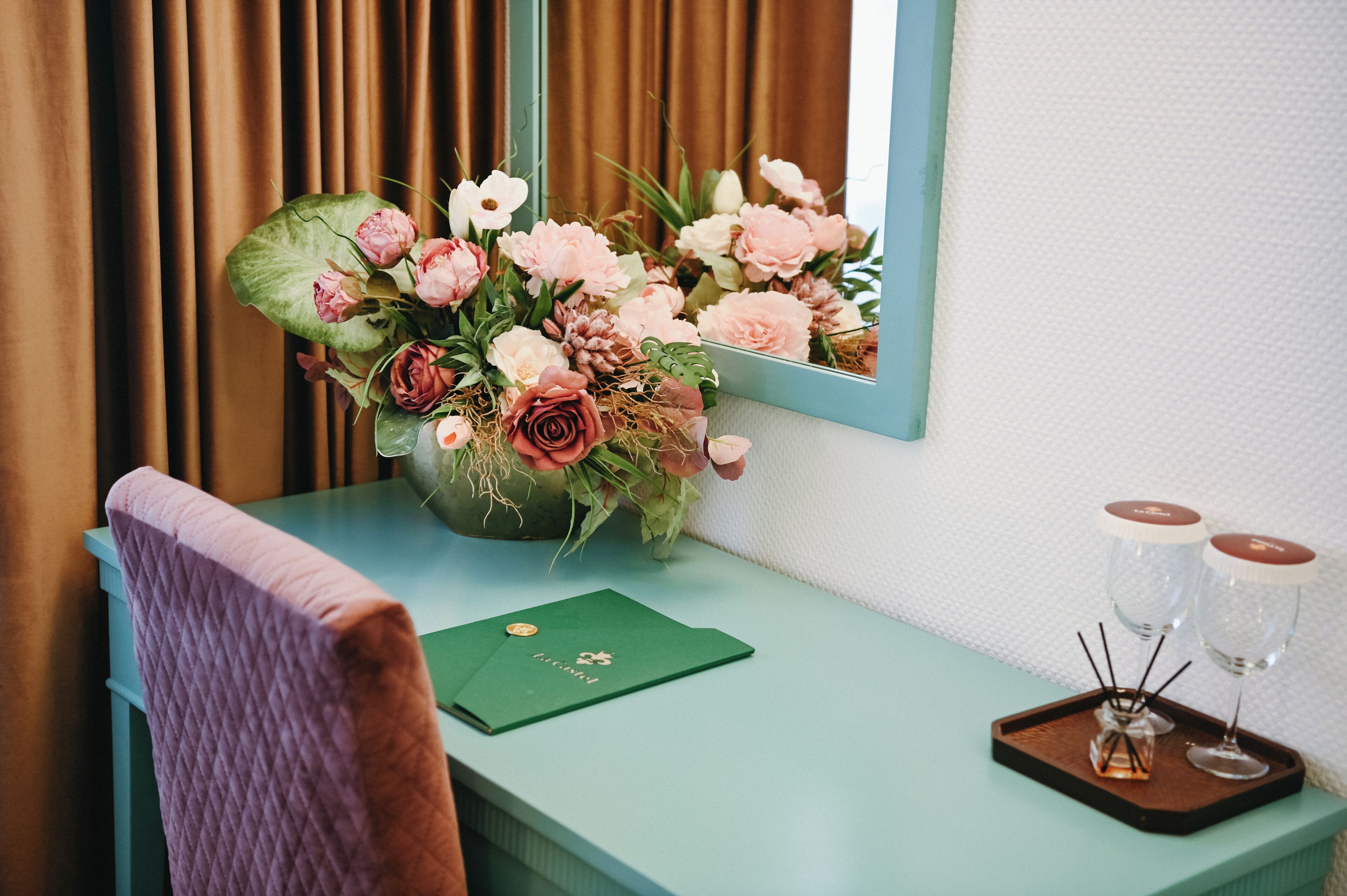 Una stanza con una scrivania verde, uno specchio, un vaso di fiori, una cartella verde e due bicchieri su un vassoio.