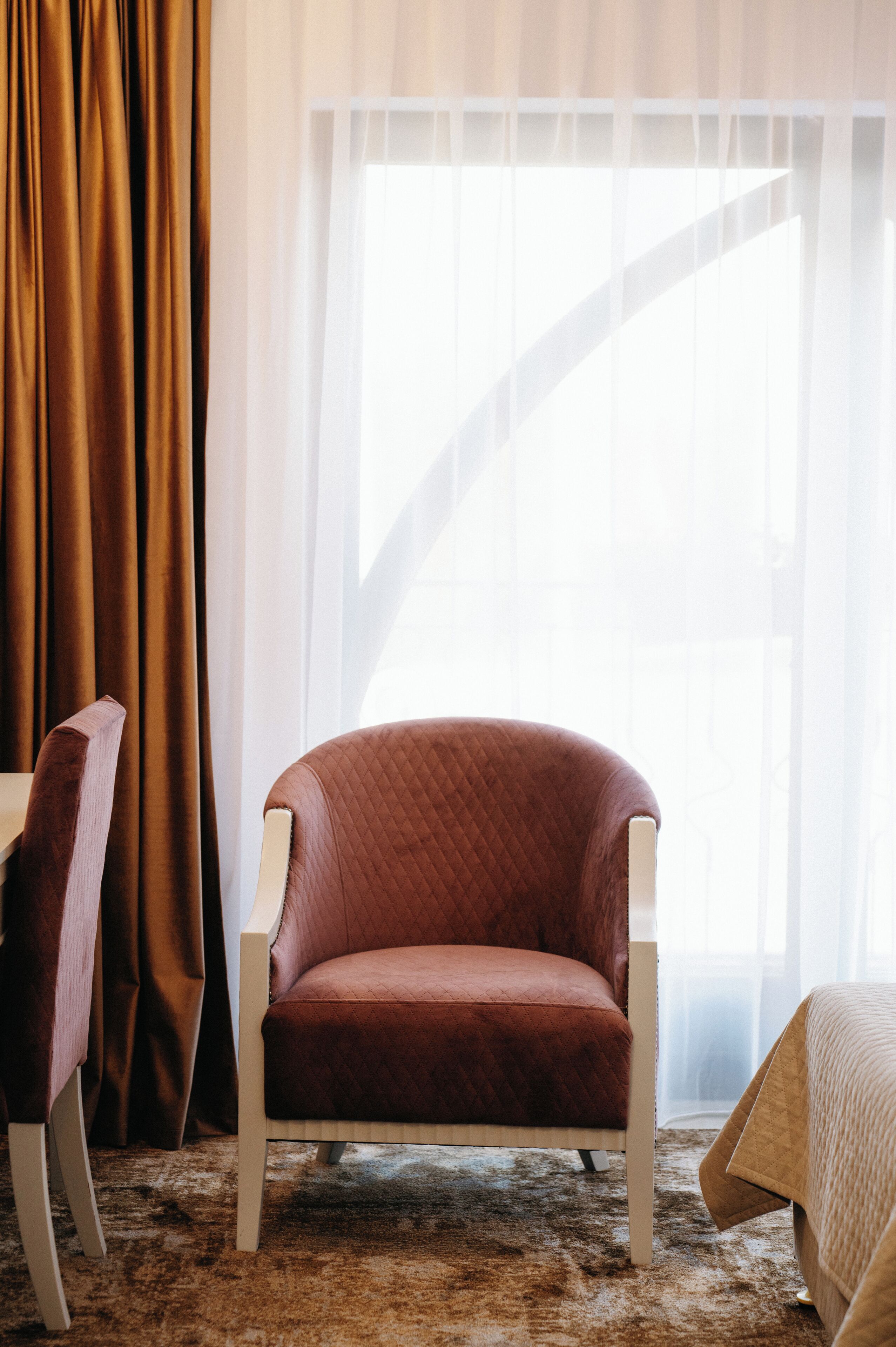 A room with a brown chair, a white chair, and a bed with a quilted cover.