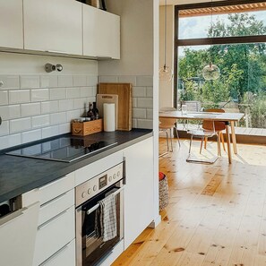 Eine moderne Küche mit weißen Schränken, einer schwarzen Arbeitsplatte und einem Backofen. Durch ein großes Fenster ist eine Essecke mit Tisch und Stühlen zu sehen.