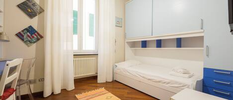 Une chambre à coucher comprenant un lit, un bureau, une chaise, un radiateur et une fenêtre avec des rideaux.