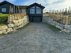 A gravel driveway leading to a dark-colored building with a stone wall on one side and a wooden fence on the other. - New cabin with views of Tisleiefjorden (Gol)