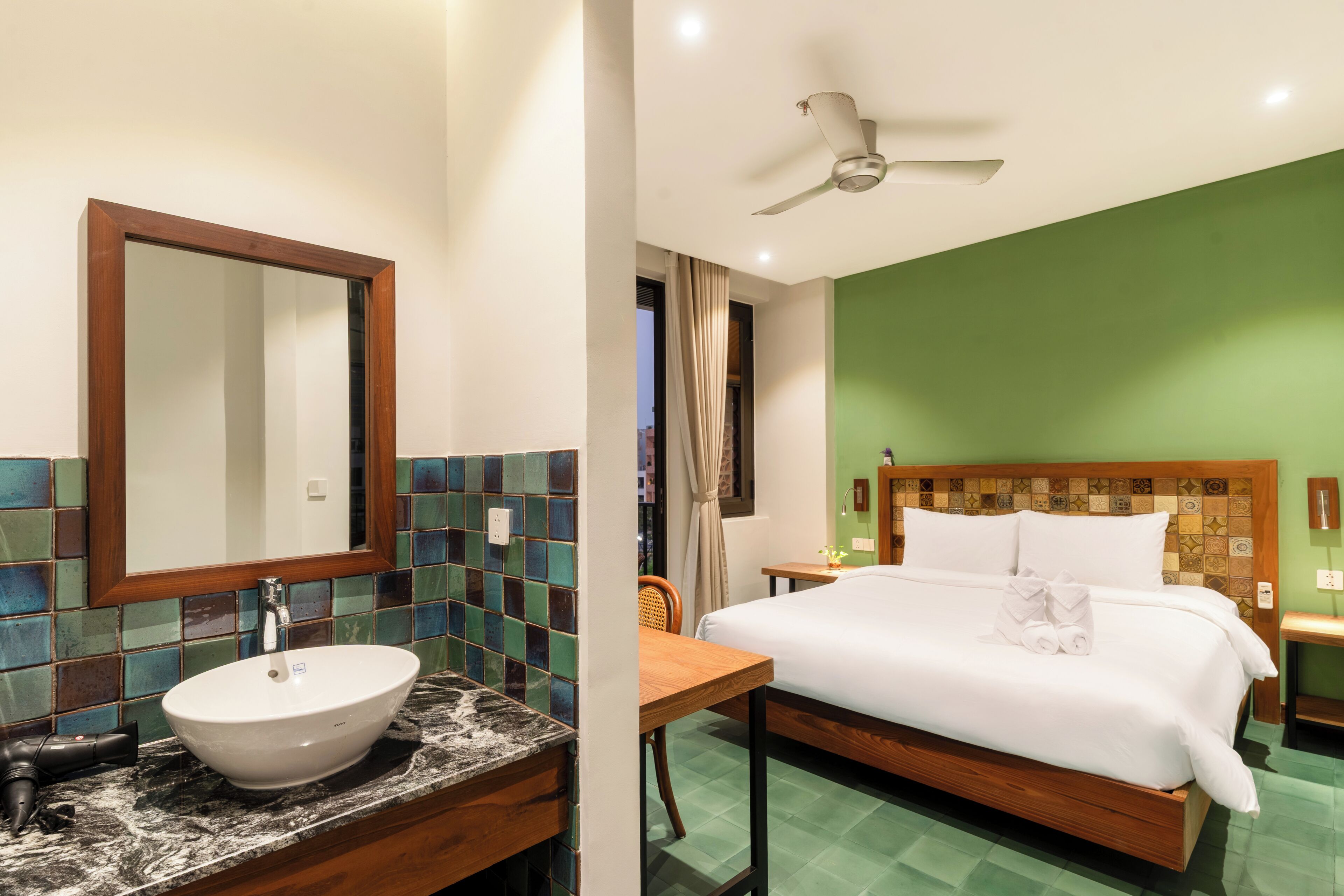 Habitación de hotel con cama, baño con lavamanos y espejo, ventilador de techo y pared verde.