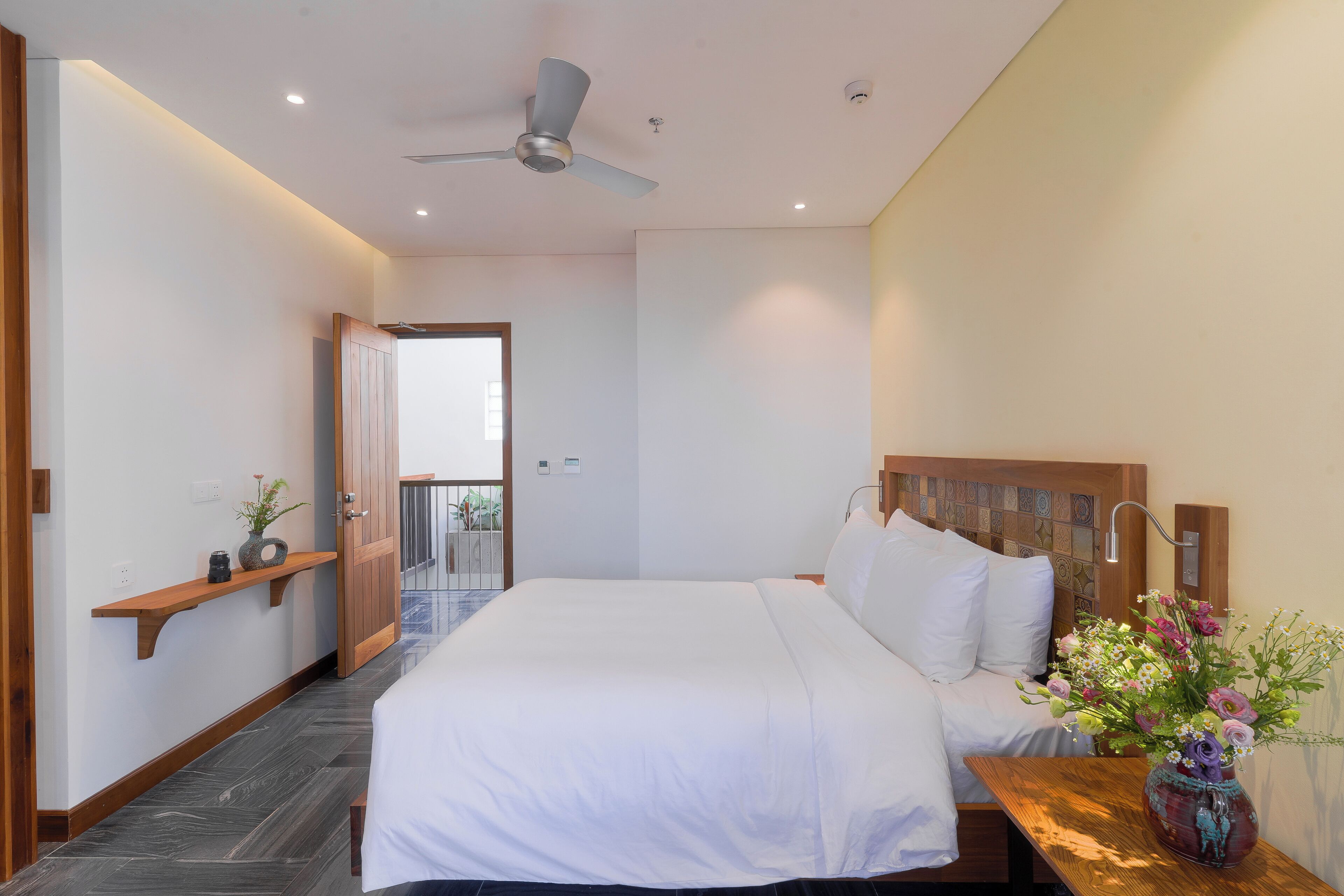 A modern hotel room with a large bed, a wooden headboard, a bedside table with a vase of flowers, and a ceiling fan.