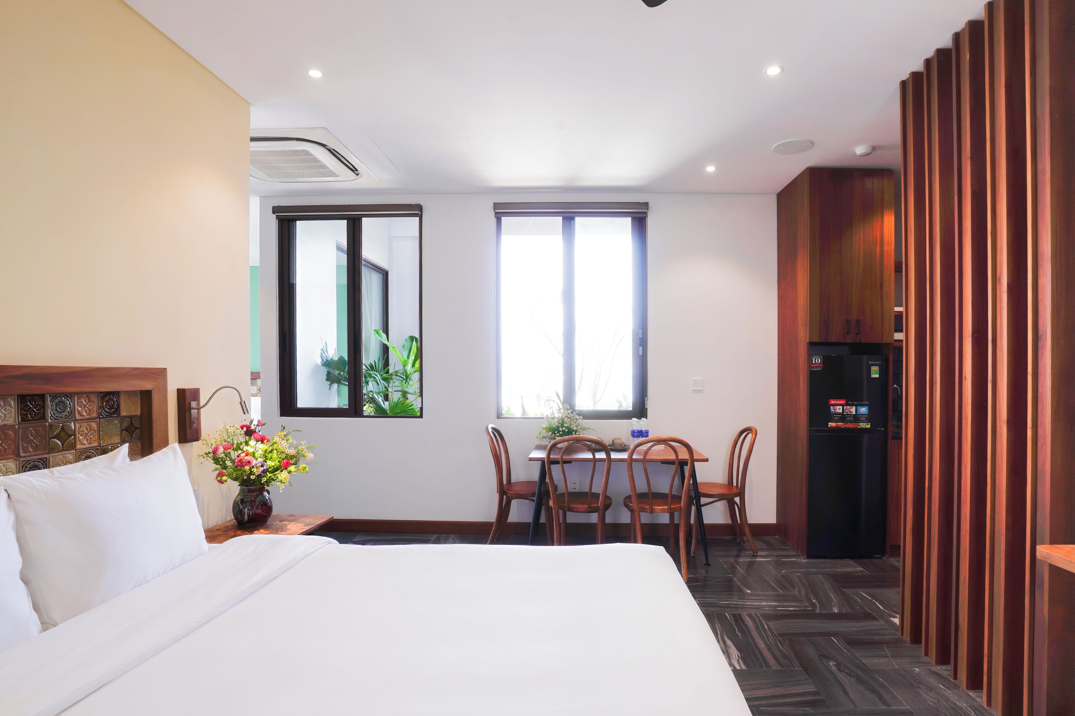 A hotel room with a bed, a dining table with chairs, a refrigerator, and a window with a view.