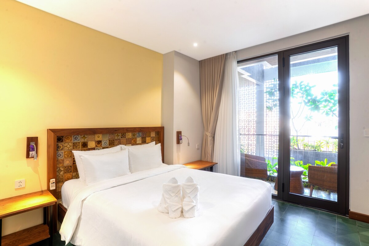 Une chambre d’hôtel avec un grand lit, une tête de lit en bois, une table de chevet, une fenêtre avec des rideaux et une vue sur l’extérieur.