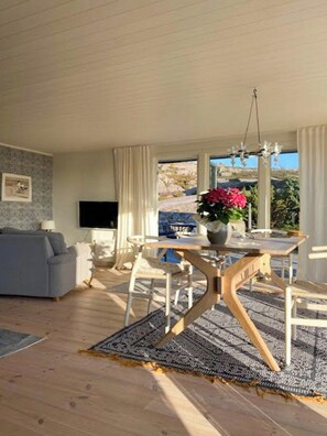 A modern living room with a grey sofa, wooden dining table, and chairs, a TV, and a large window with a view. - Seaside house with pool near Hällevikstrand (Ellös)