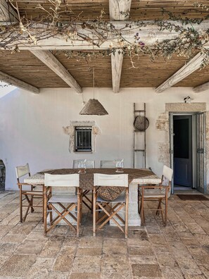 Un espace de restauration extérieur de style rustique, avec un sol carrelé en pierre, des chaises en bois, une nappe à motifs et un luminaire suspendu.