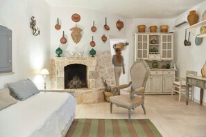 Ein Zimmer mit Kamin aus Stein, einem Bett, einem Sessel, einem Esstisch und einer Küchenzeile mit Schränken und hängenden Töpfen.