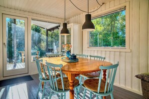 Een eethoek met een houten tafel en stoelen, hanglampen en grote ramen met uitzicht op bomen.