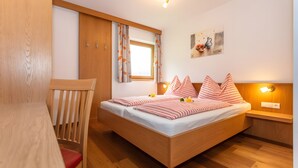 Un dormitorio con una cama y una silla de madera, un armario, un ventanal con cortinas, una lámpara de pared y un cuadro.