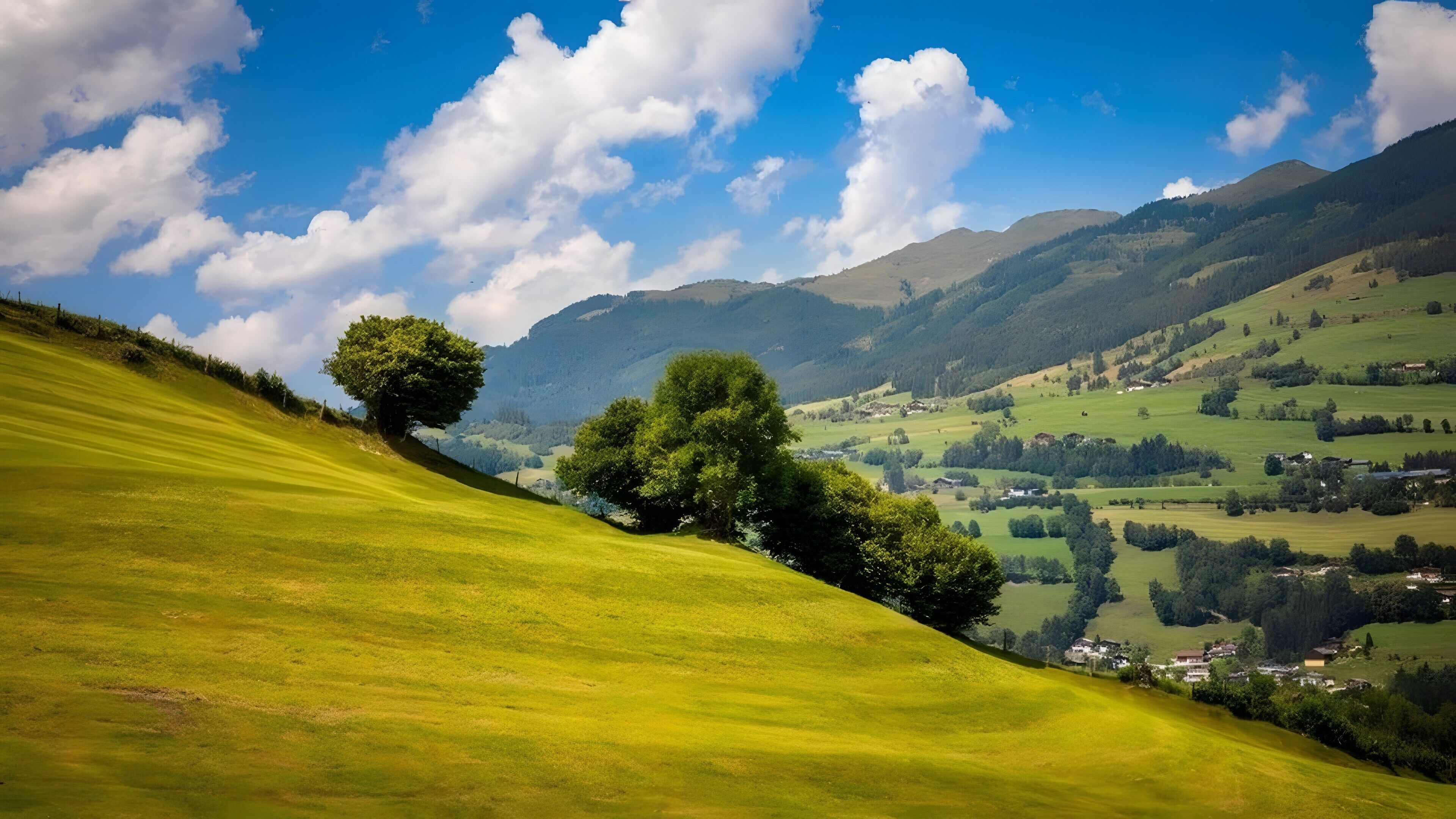 A hilly landscape with a prominent tree on a slope, a valley with scattered houses, and distant mountains under a clear sky.