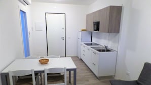 Una cucina compatta con mobili bianchi, forno a microonde integrato e lavello. Nell'area soggiorno, un tavolo da pranzo con sedie e un divano grigio.
