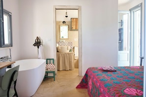 Ein Zimmer mit Badewanne, einem Bett mit floralem Überwurf, einem Sessel und Blick ins Badezimmer.