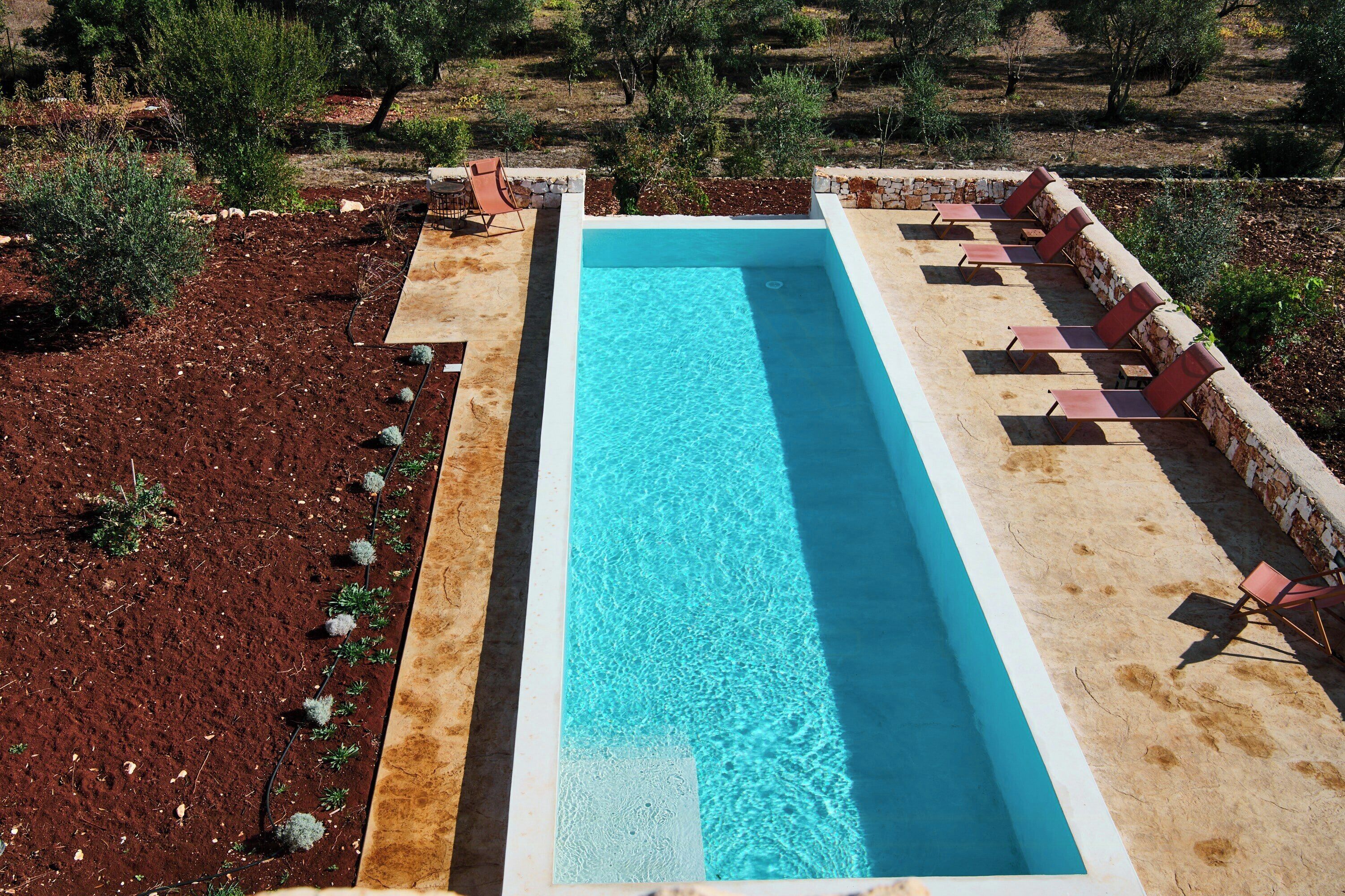 Una piscina rectangular con superficie de agua azul cristalina, rodeada por una terraza de piedra y sillas de descanso rojas.