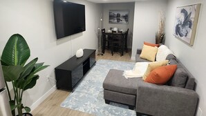 En moderne stue med et fladskærms-tv, en grå sofa og et spiseområde med bord og stole.