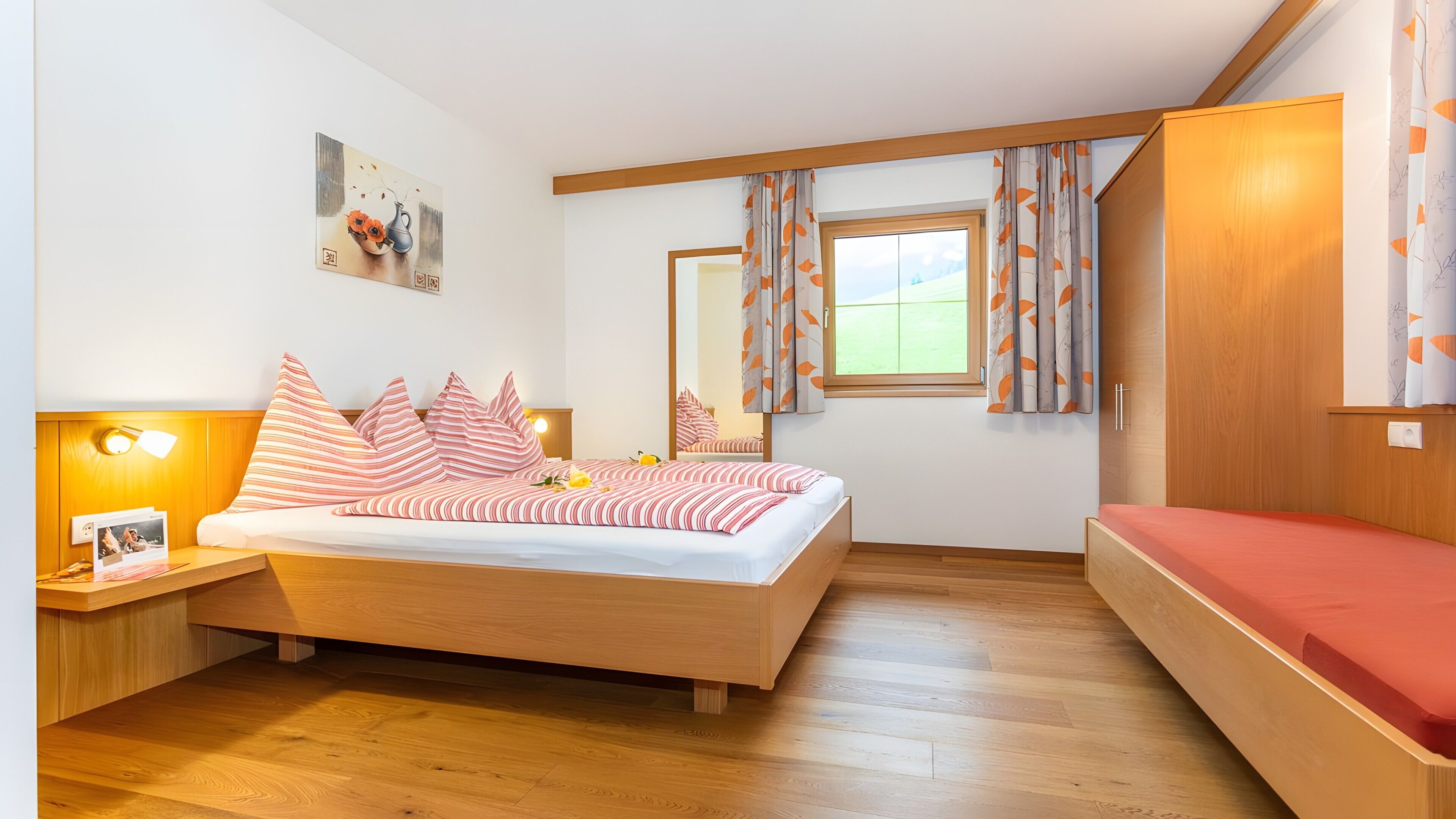 Une chambre à coucher comprenant un lit, une table de chevet, un miroir, une fenêtre avec des rideaux et une armoire en bois.